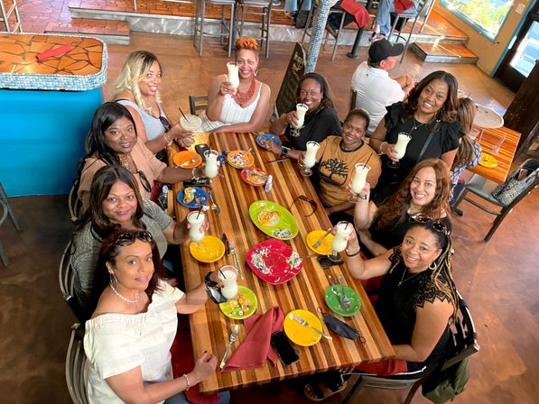 Group of women toasting with frozen cocktails around a long striped wooden table in a bright casual restaurant, colorful plates and napkins creating a festive brunch scene.