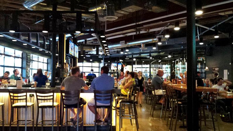 Bustling industrial-style brewery taproom with a long illuminated bar, patrons on bar stools, exposed ductwork, visible brewing tanks and wall-mounted TVs.