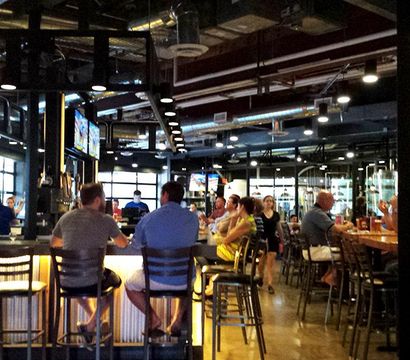 Bustling industrial-style brewery taproom with a long illuminated bar, patrons on bar stools, exposed ductwork, visible brewing tanks and wall-mounted TVs.
