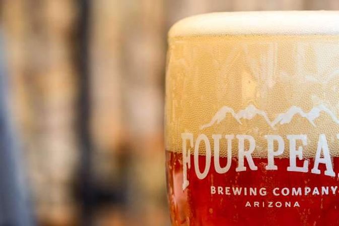 Close-up of amber craft beer with a creamy head in a branded pint glass showing a mountain logo and 'Arizona' text, blurred brewery background