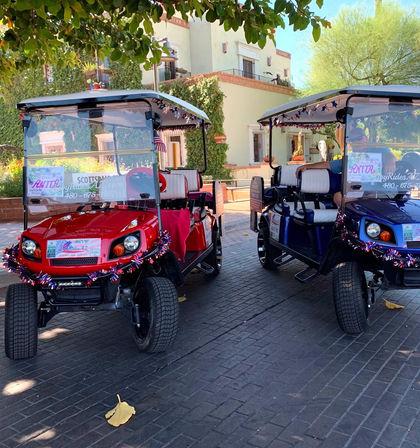 Two festive red and blue golf carts with star-spangled tinsel parked under trees in a sunny Scottsdale courtyard with Spanish-style buildings