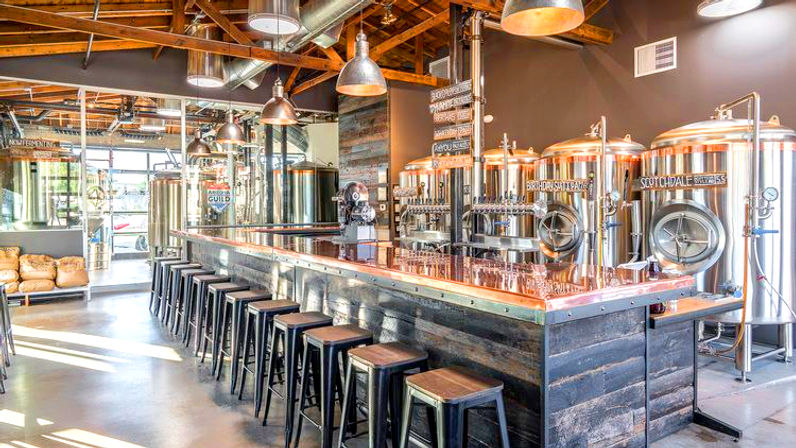 Sunlit industrial-style craft brewery taproom with a long copper-topped bar, row of black metal stools, shiny stainless-steel fermentation tanks, exposed wooden beams and hanging pendant lights.