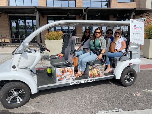 Four smiling women seated in a white open-air tour cart on a sunny Arizona city street, promotional panels for wine-tasting and taco tours visible on the vehicle