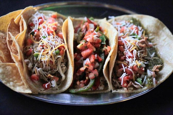 Three colorful street-style corn tortilla tacos on a metal plate filled with shredded chicken, pico de gallo, sautéed peppers, shredded cheese and green salsa.