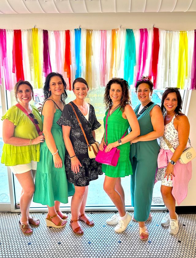 Six women in colorful summer outfits and sandals smiling and posing inside a bright storefront, standing in front of a rainbow tassel backdrop hanging across the window.