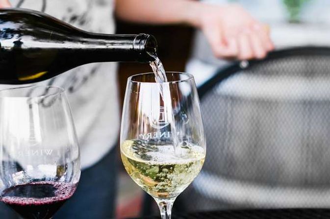 Close-up of white wine being poured into a stemmed glass on a sunny patio, with a second glass of red wine nearby — casual wine-tasting moment.