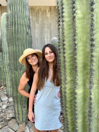 Two smiling women posing between tall saguaro cacti in a sunlit Southwestern desert garden — one wearing a straw hat and black dress, the other in a light blue dotted summer dress.