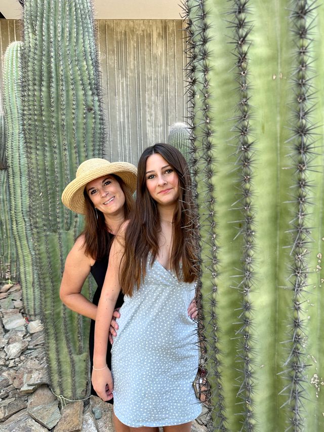 Two smiling women posing between tall saguaro cacti in a sunlit Southwestern desert garden — one wearing a straw hat and black dress, the other in a light blue dotted summer dress.
