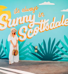 Person in white dress and sunhat holding a round straw bag, posing in front of a bright turquoise mural reading “It’s always Sunny in Scottsdale” with stylized agave plants — colorful Scottsdale street art.