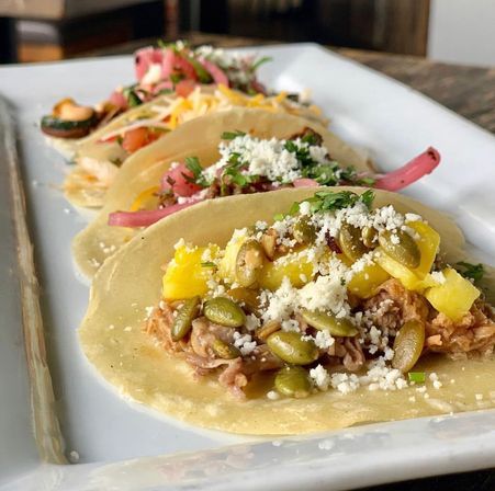 Close-up of three Mexican-style corn tortilla tacos on a white rectangular plate featuring shredded pork with pineapple, toasted pepitas, crumbled cotija, pickled red onions and cilantro on a wooden table