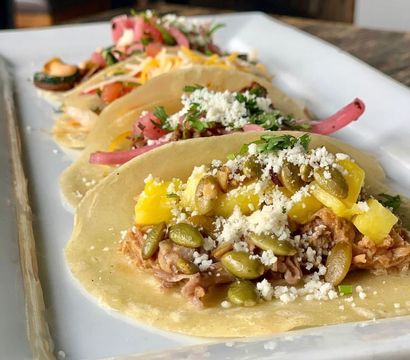 Close-up of three Mexican-style corn tortilla tacos on a white rectangular plate featuring shredded pork with pineapple, toasted pepitas, crumbled cotija, pickled red onions and cilantro on a wooden table