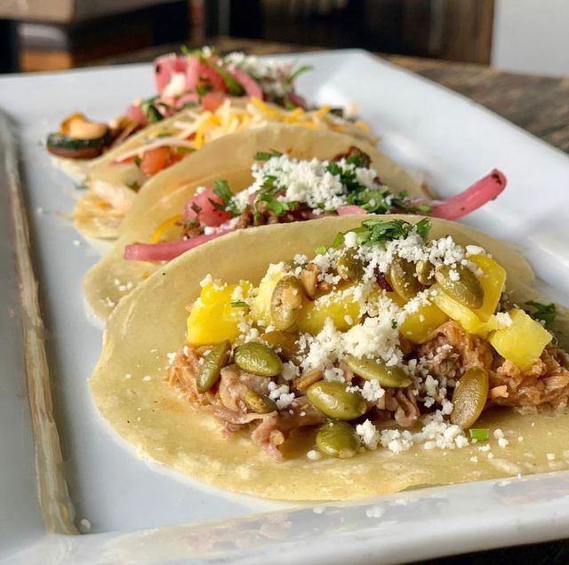 Close-up of three Mexican-style corn tortilla tacos on a white rectangular plate featuring shredded pork with pineapple, toasted pepitas, crumbled cotija, pickled red onions and cilantro on a wooden table