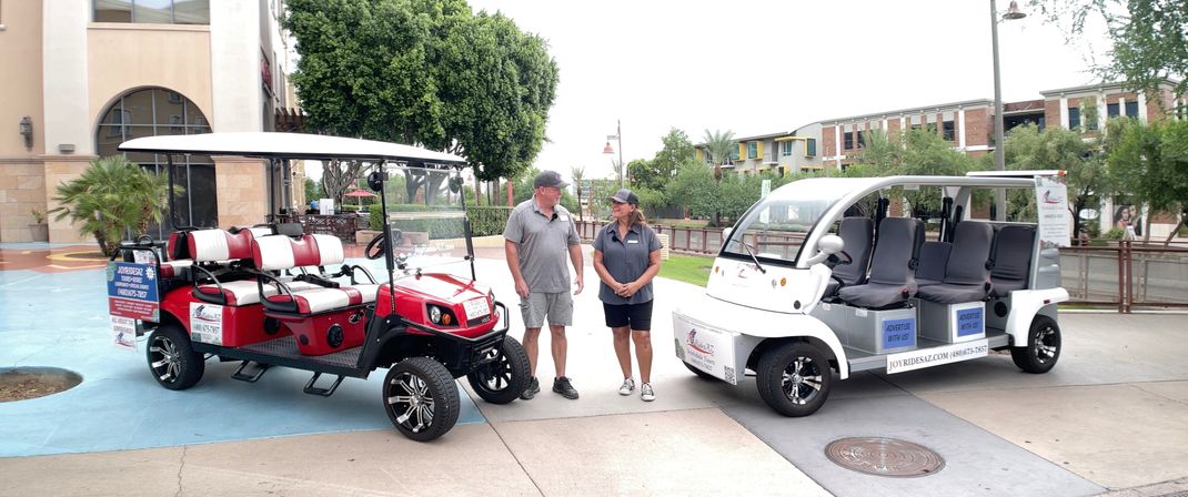Two staff members stand between a red and white open-air electric shuttle cart parked on a sunny outdoor shopping plaza with trees and low-rise mixed-use buildings.