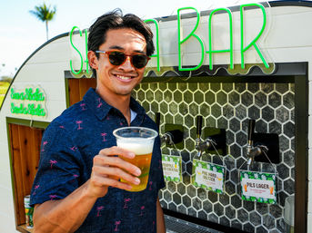 Smiling man in sunglasses holding a plastic beer cup in front of an outdoor beach-style bar trailer with a neon BAR sign, three taps and a palm tree in the background.