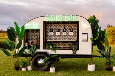White mobile bar trailer with glowing neon sign, four drink taps and bottles against hex tile, surrounded by potted tropical plants on a grassy field at sunset.