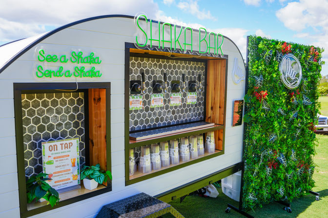 Outdoor surf‑themed mobile bar trailer with neon signage, four drink taps, stacked cups, on‑tap menu and a lush vertical plant wall on a sunny park lawn