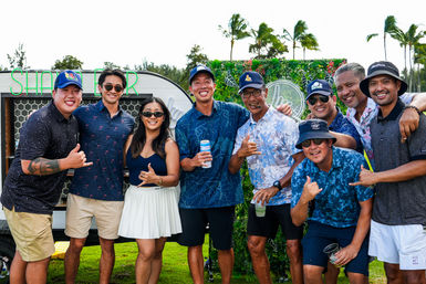 Nine people in casual island-style shirts and hats posing on a grassy tropical lawn with palm trees and a neon food-truck backdrop, smiling and flashing shaka signs while holding drinks.