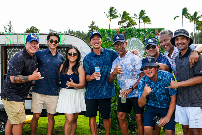 Nine people in casual island-style shirts and hats posing on a grassy tropical lawn with palm trees and a neon food-truck backdrop, smiling and flashing shaka signs while holding drinks.