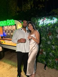 Smiling couple in evening attire posing at a tropical outdoor party by a neon-lit tiki drink cart and a leafy photo wall with a custom neon sign.