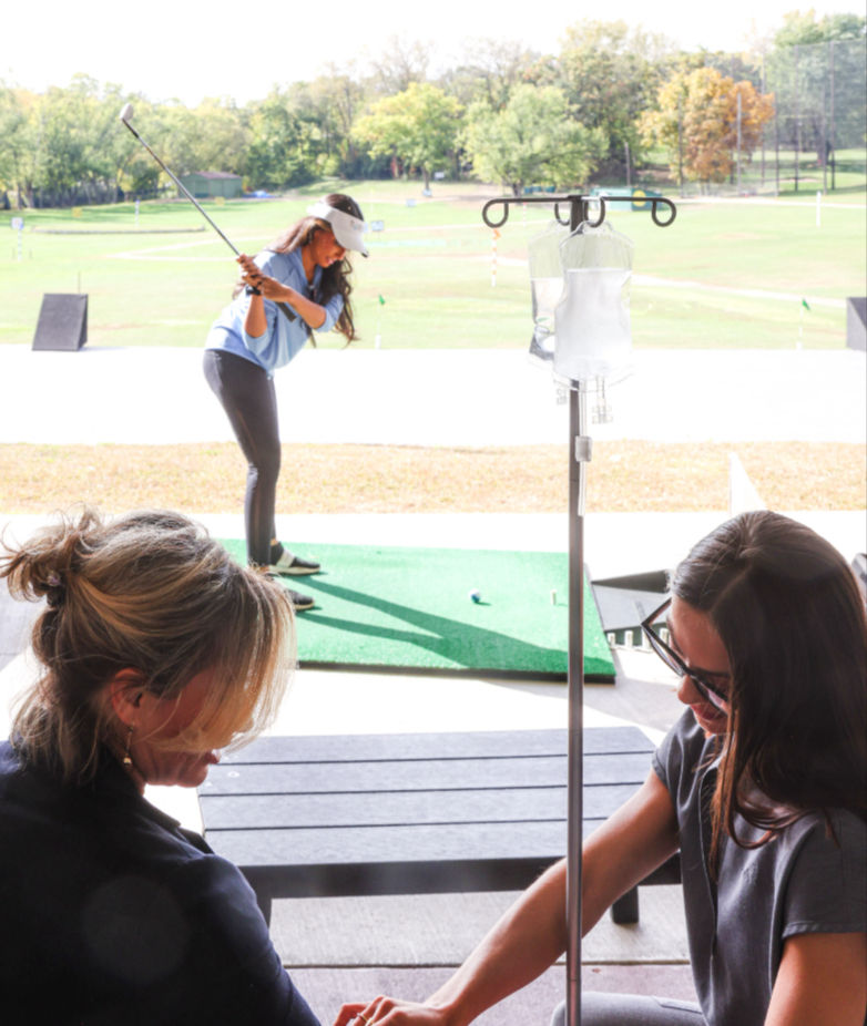 Golfer mid-swing on an outdoor driving range while two people chat at a bench in the foreground beside an IV pole with a clear bag.