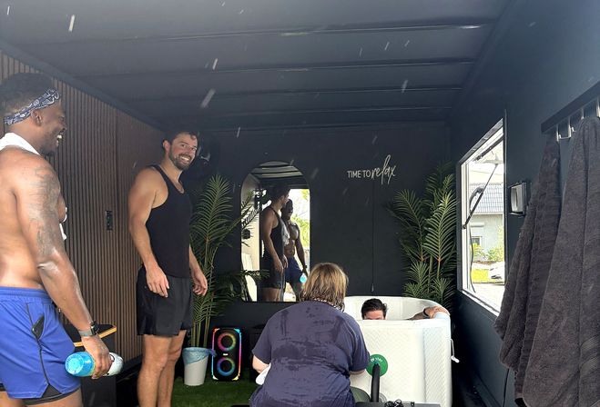Group of men laughing around a portable cold-plunge tub inside a dark wellness trailer with potted plants, mirror reflection, colorful speaker and a wall sign reading Time to relax.