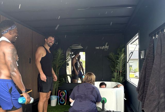 Group of men laughing around a portable cold-plunge tub inside a dark wellness trailer with potted plants, mirror reflection, colorful speaker and a wall sign reading Time to relax.