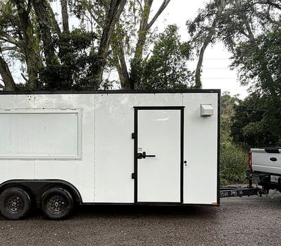 White twin-axle mobile sauna trailer with side door and closed service window, hitched to a white pickup truck and parked on wet pavement beneath large trees along a tree-lined road.