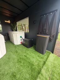 Backyard spa cabana with inflatable white hot tub on green artificial turf, black heater unit, three dark towels on hooks, neon "Time to relax" sign and trees visible through the window.