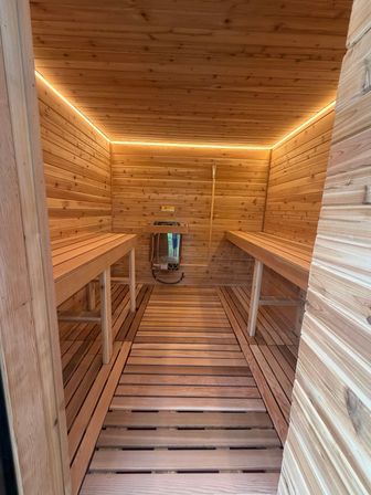 Modern cedar sauna interior with parallel wooden benches, slatted floor, wall-mounted heater and warm LED cove lighting.