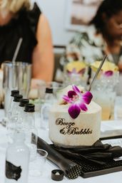 Tropical coconut cocktail engraved with "Booze & Bubbles", topped with a purple orchid and metal straw, surrounded by mini liquor bottles and bar tools on a white event table.