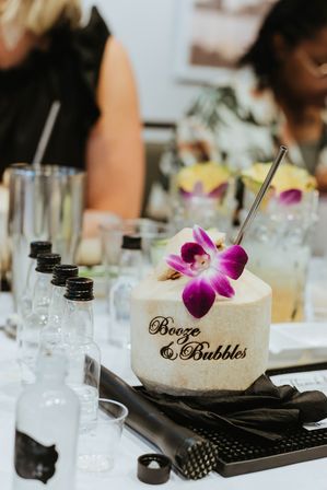 Tropical coconut cocktail engraved with "Booze & Bubbles", topped with a purple orchid and metal straw, surrounded by mini liquor bottles and bar tools on a white event table.