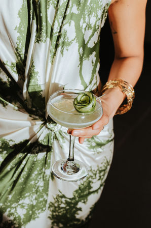 Chic cucumber-topped pale green cocktail in a coupe glass held by a person in a white-and-green patterned dress with stacked gold bangles.