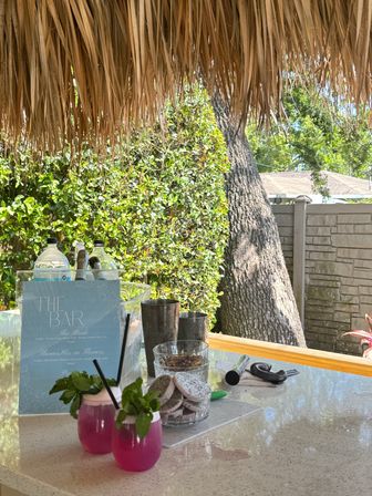 Tropical backyard tiki bar under a thatched roof with two bright pink, mint‑garnished cocktails on the counter, bar menu, bottles and leafy hedge in the background.