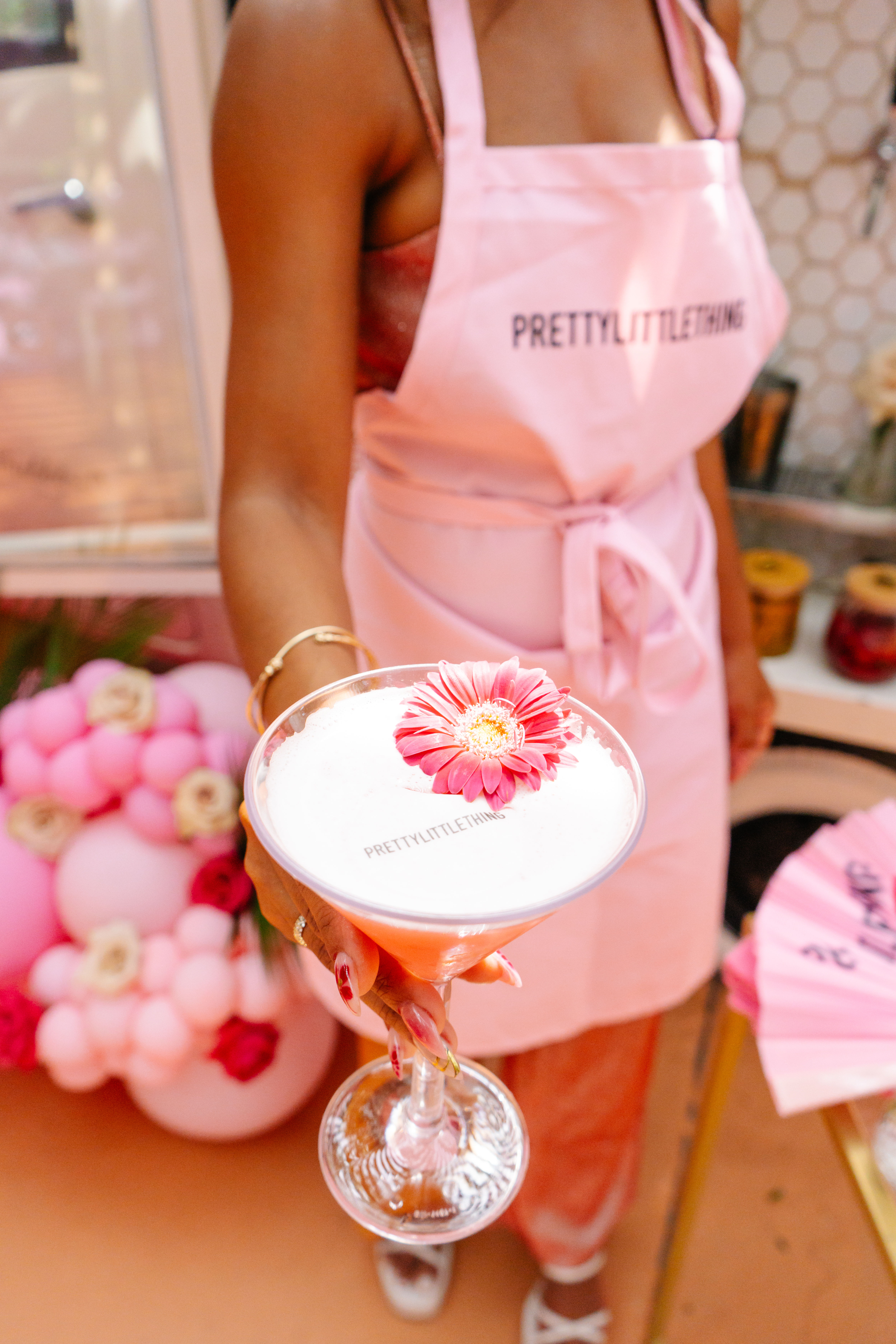 Hand-held frothy pink cocktail topped with a pink gerbera flower, offered by a person in a pink apron with pastel pink balloon decor and party setup in the background.