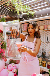 Woman in a pink apron pouring a pink cocktail from a shaker into a martini glass at a sunny outdoor patio pop-up bar decorated with pink balloons, roses, and tropical greenery.