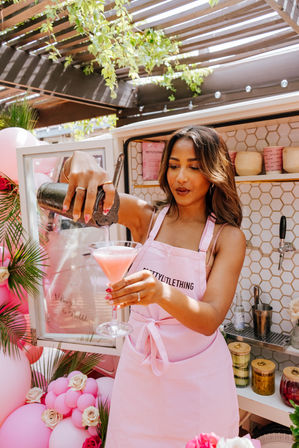 Woman in a pink apron pouring a pink cocktail from a shaker into a martini glass at a sunny outdoor patio pop-up bar decorated with pink balloons, roses, and tropical greenery.