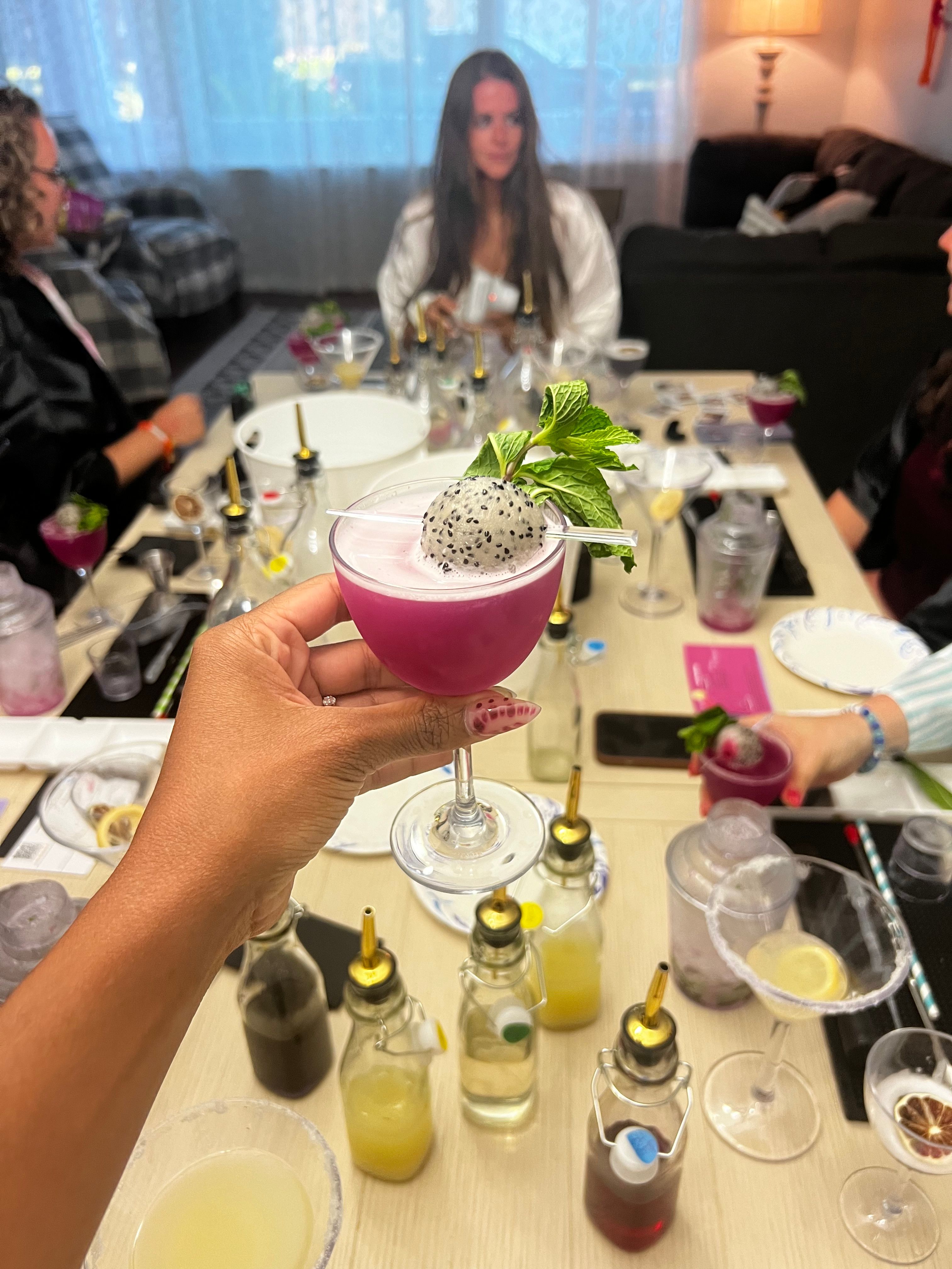 Hand holding vibrant magenta dragon-fruit cocktail in a coupe glass, topped with a dragonfruit ball and mint, surrounded by bottles and tools on a home mixology party table with blurred guests in a living room.