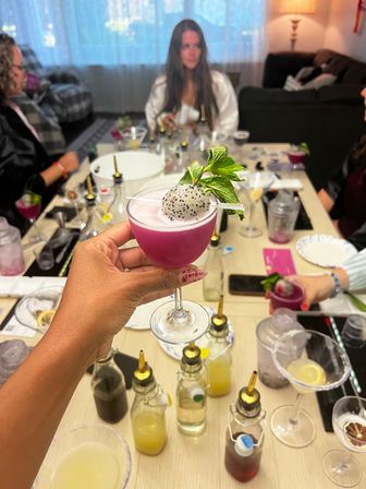 Hand holding vibrant magenta dragon-fruit cocktail in a coupe glass, topped with a dragonfruit ball and mint, surrounded by bottles and tools on a home mixology party table with blurred guests in a living room.