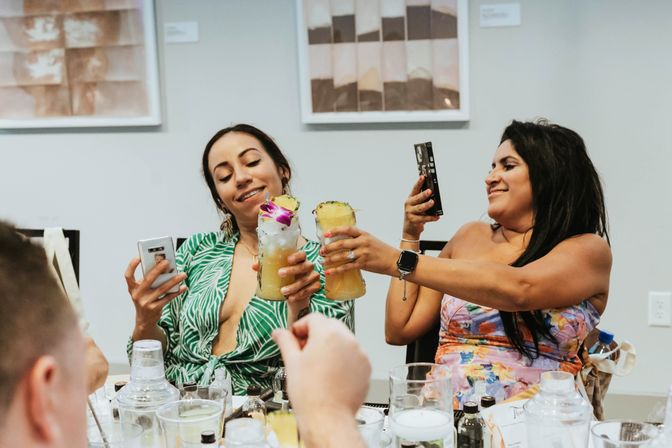 Two friends clinking pineapple-topped tropical cocktails with orchid garnish at an indoor brunch table, snapping photos with smartphones