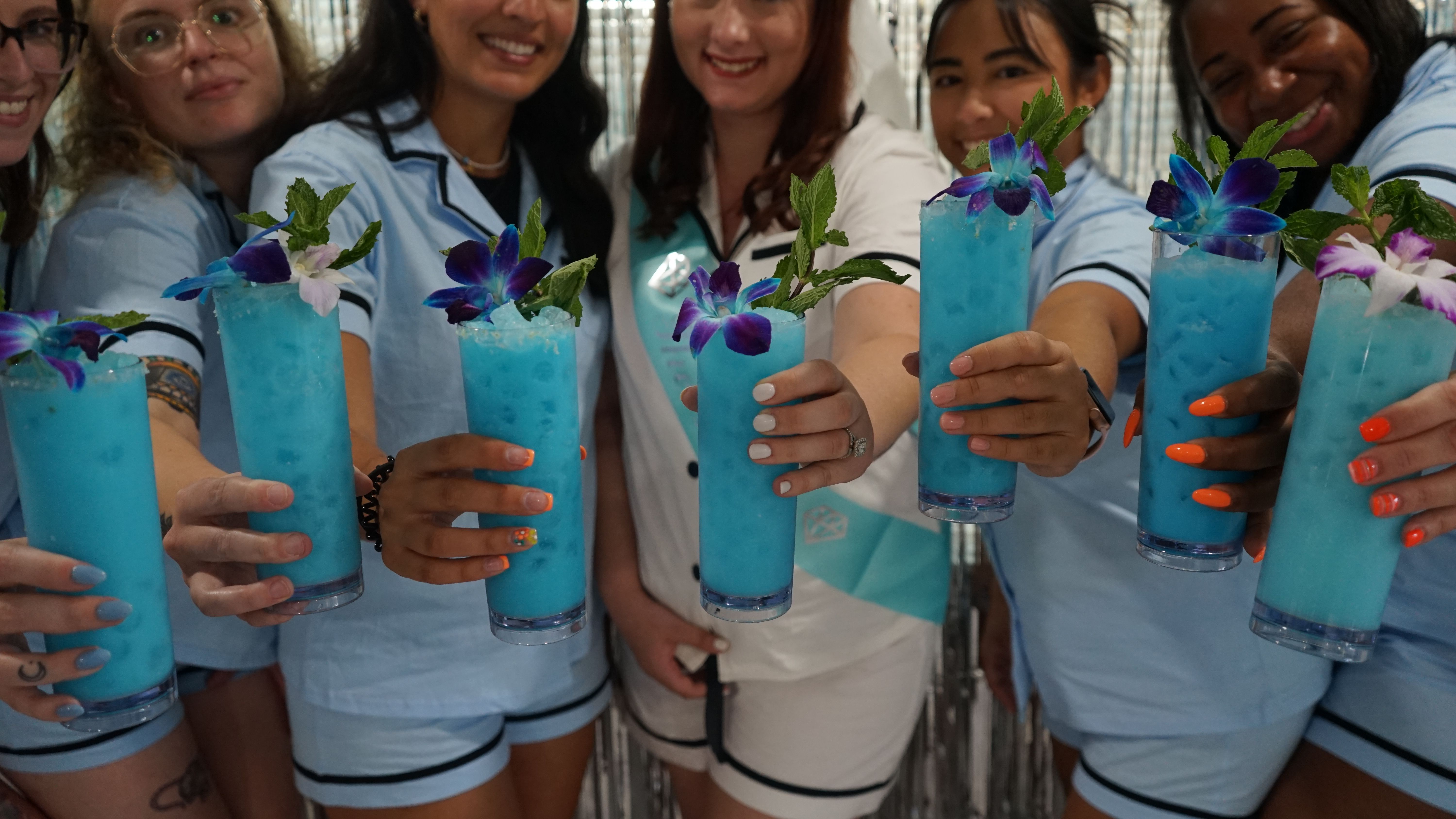 Friends in matching light-blue outfits toasting tall icy turquoise cocktails garnished with purple orchids and mint, close-up of hands with colorful nails.