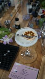 Lavender gin sour in a coupe glass with frothy white foam, garnished with a dried orange wheel and lavender sprig, sitting on a wooden bar counter among cocktail tools and a recipe card.