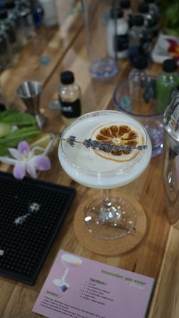 Lavender gin sour in a coupe glass with frothy white foam, garnished with a dried orange wheel and lavender sprig, sitting on a wooden bar counter among cocktail tools and a recipe card.