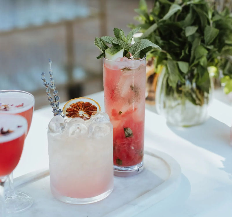 Three pink summer cocktails on a white marble tray — a crushed-ice lavender drink with a dried orange wheel, a tall minty pink highball with cucumber and mint, and a coupe glass, with fresh mint in a vase behind.
