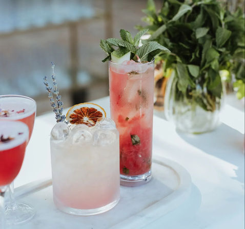 Three pink summer cocktails on a white marble tray — a crushed-ice lavender drink with a dried orange wheel, a tall minty pink highball with cucumber and mint, and a coupe glass, with fresh mint in a vase behind.