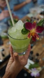 Hand holding a bright green tropical cocktail with crushed ice, cucumber ribbon and purple orchid garnish at a lively bar