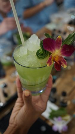 Hand holding a bright green tropical cocktail with crushed ice, cucumber ribbon and purple orchid garnish at a lively bar