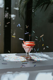 Sunlit pink milkshake in a glass with a sprinkle-coated rim, topped with a stacked pink donut and a lit sparkler, surrounded by rainbow confetti on a white table with palm leaf shadows.