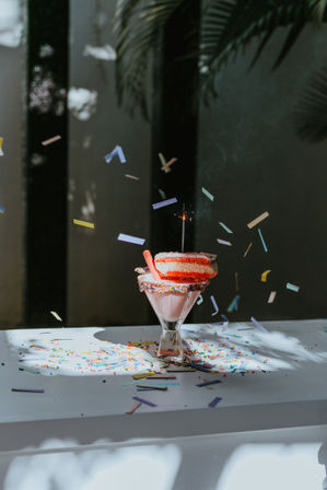 Sunlit pink milkshake in a glass with a sprinkle-coated rim, topped with a stacked pink donut and a lit sparkler, surrounded by rainbow confetti on a white table with palm leaf shadows.