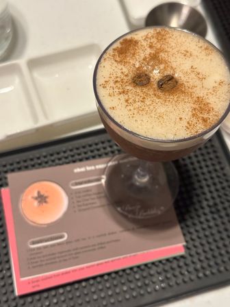 Creamy espresso martini in a coupe glass topped with cinnamon and two coffee beans, sitting on a black bar mat over a blurred recipe card.