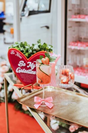Pink Valentine-themed cocktail in a stemmed glass garnished with a strawberry heart and mint, set on a brass tray with a pink bow and a blurred heart-shaped Be My Galentine sign in a boutique cafe display.